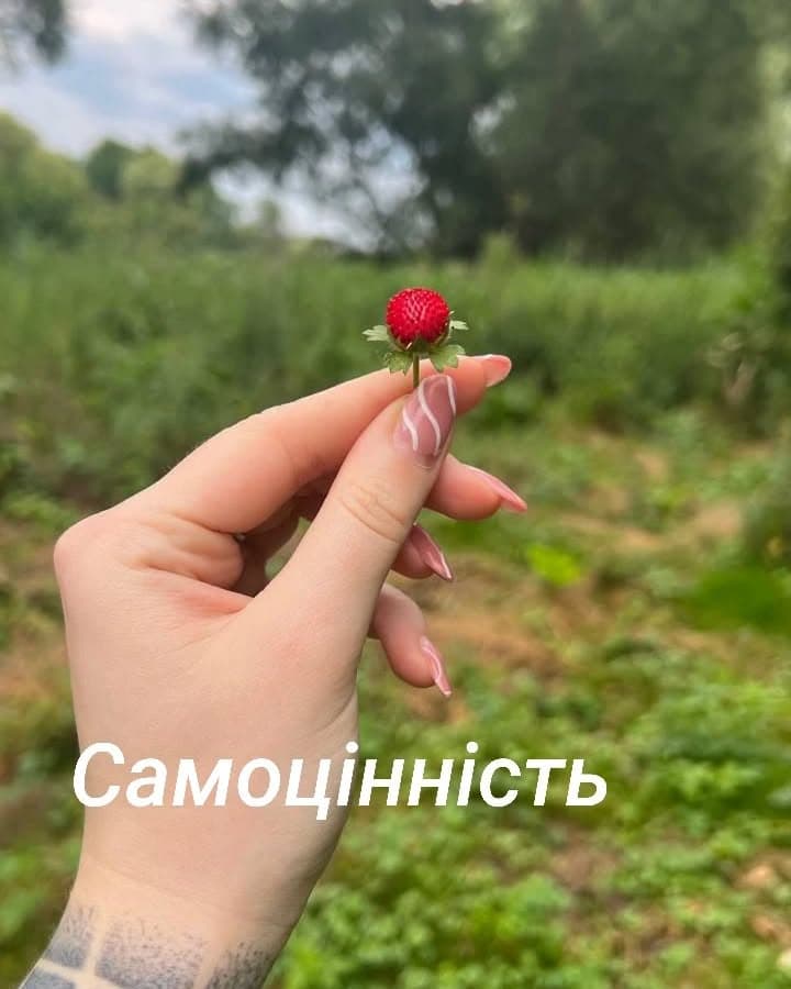 Рука з довгим манікюром тримає маленьку суницю на тлі зеленої галявини; напис «Самоцінність»