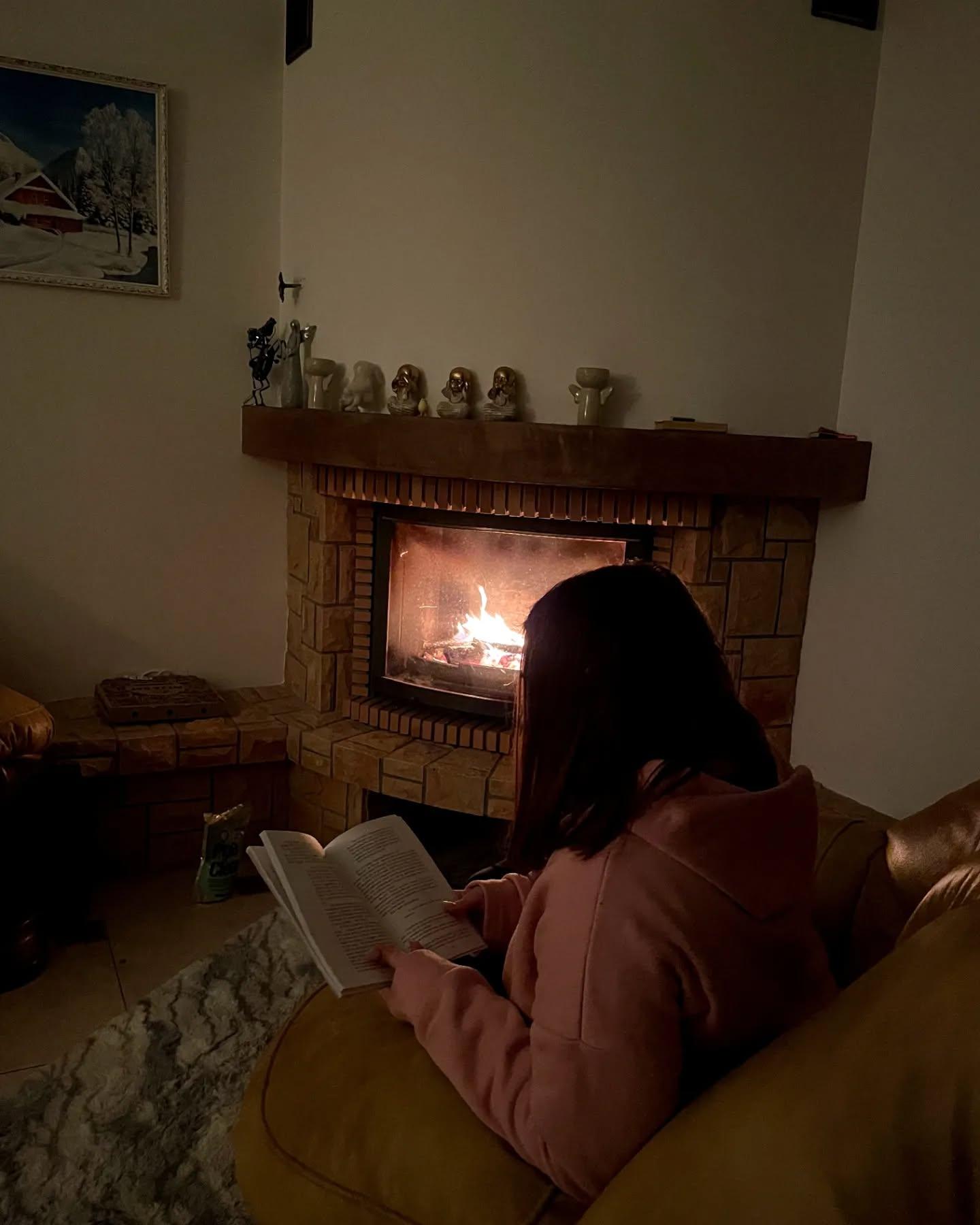Дівчина в рожевому худі читає книжку в кріслі біля каміна