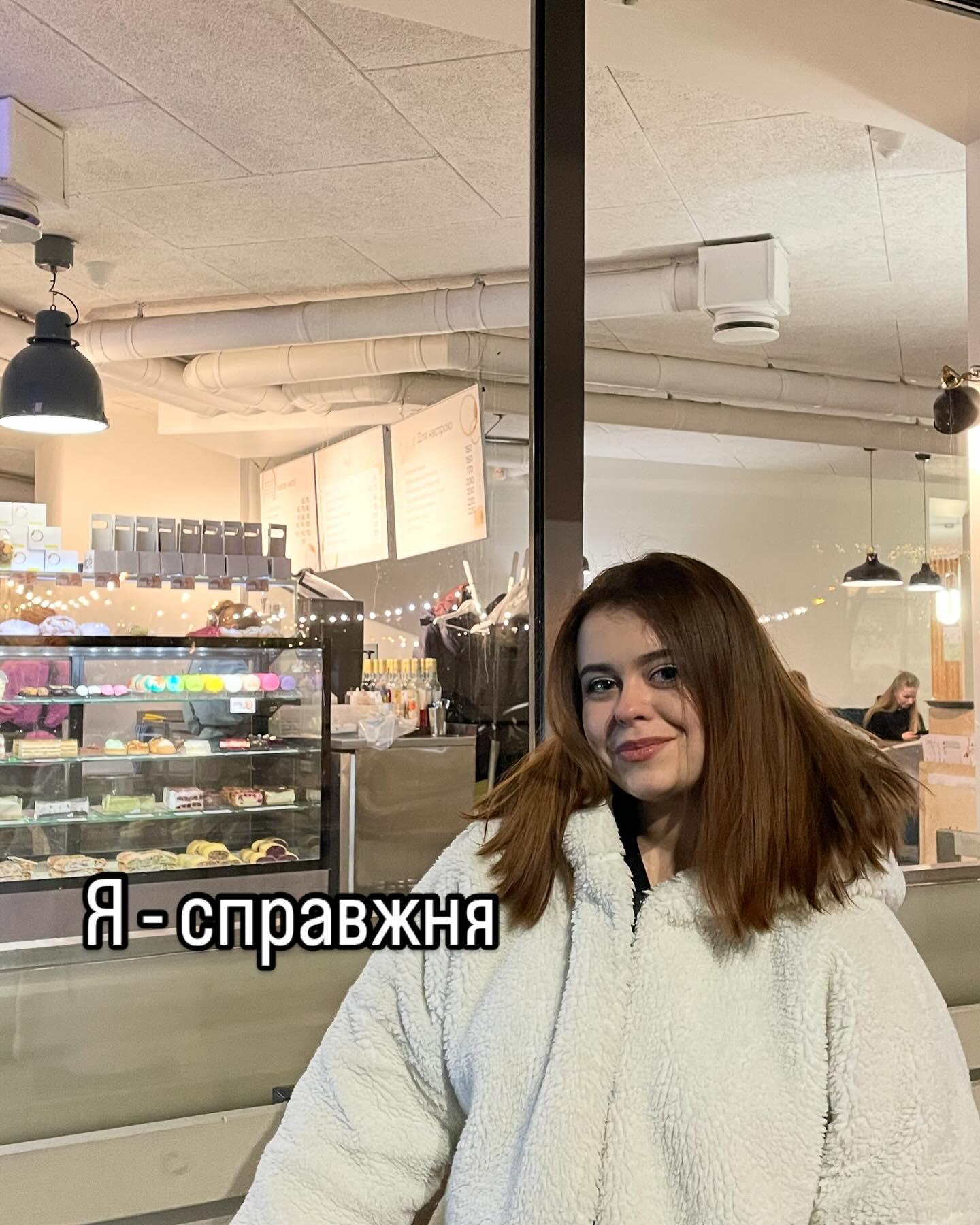 Марія Дерев'янченко в білій пухнастій куртці у світлому кафе з вітринами; напис «Я - справжня»